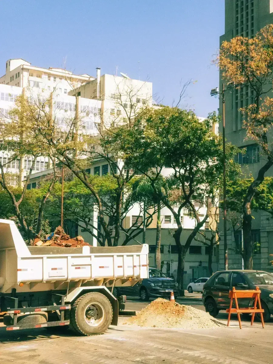 Caminhão caçamba removendo entulho em rua urbana, com pilhas de terra, cone, carros e prédios ao fundo